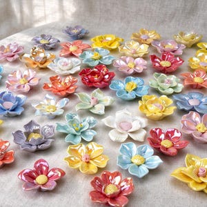 May include: A collection of small, individual ceramic flowers in various pastel and vibrant colors including yellow, pink, blue, and orange. Each flower features layered petals and a textured, raised center, arranged in a scattered pattern.