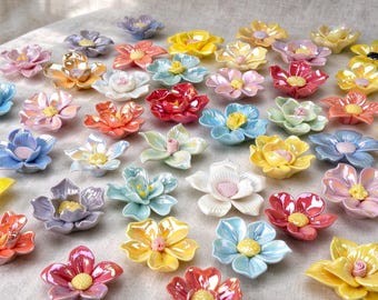 May include: A collection of small, individual ceramic flowers in various pastel and vibrant colors including yellow, pink, blue, and orange. Each flower features layered petals and a textured, raised center, arranged in a scattered pattern.