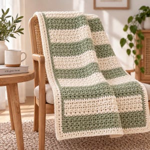 Könnte beinhalten: Eine gehäkelte Decke mit abwechselnden horizontalen Streifen aus salbeigrünem und cremefarbenem Garn. Das Textil weist ein strukturiertes, sich wiederholendes Stichmuster auf und ist an allen vier Kanten mit einem cremefarbenen Rand versehen.