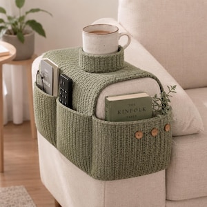 May include: A sage green knitted sofa armrest organizer featuring a built-in cup holder, multiple side pockets for storage, and three decorative wooden buttons. The organizer holds a ceramic mug, a remote control, and a book titled The Kinfolk Table.