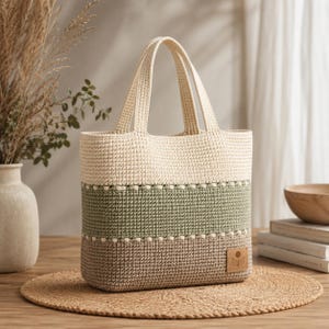 Könnte beinhalten: Eine Häkeltasche mit einem Color-Block-Design aus horizontalen Streifen in Creme, Salbeigrün und Hellbraun. Die Tasche hat zwei passende cremefarbene Henkel und einen kleinen quadratischen Aufnäher in Lederoptik mit einem Blumen-Symbol.
