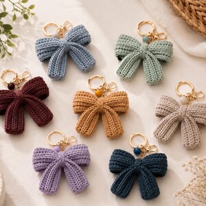 Può includere: Sette portachiavi a forma di fiocco all'uncinetto in vari colori, tra cui blu, verde salvia, bordeaux, giallo senape, beige, lavanda e blu scuro. Ogni fiocco presenta un anello in metallo dorato con una piccola perlina abbinata.