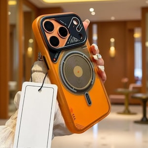 Può includere: Una custodia per smartphone arancione con un modulo fotocamera nero a tre lenti circolari, un anello di ricarica magnetico centrale e una piccola etichetta rettangolare attaccata con un cordino nero. La custodia ha una finitura arancione.