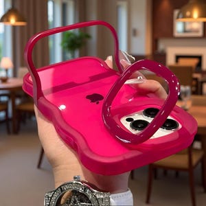 Puede incluir: Una funda de teléfono rosa brillante y translúcida con un asa integrada y un soporte de anillo circular. La funda está diseñada para un smartphone con un sistema de cámara de tres lentes, mostrando el logotipo de Apple a través del material.