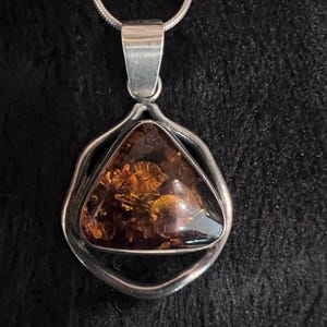 May include: A triangular amber pendant set in a smooth silver frame, suspended from a thin silver snake chain. The amber displays deep orange and brown tones with visible internal inclusions, held by a polished silver bail at the top.