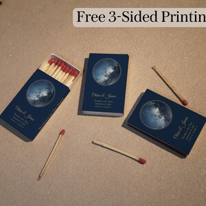 May include: Three dark blue matchboxes with a circular galaxy graphic, gold text reading Olivia & James, Written in the Stars, and a date. One box is open showing wooden matches with red tips, while two loose matches lie on the surface nearby.
