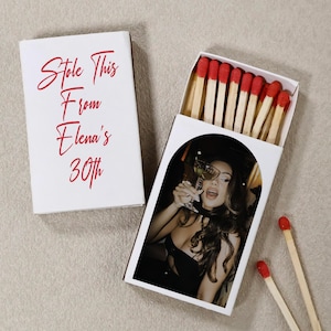 May include: A white matchbox with the red text Stole This From Elena's 30th. The sliding tray features a photograph of a person holding a glass, and the box contains wooden matches with red tips. Two additional matches with red tips lie nearby.
