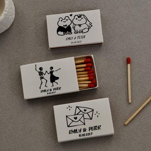 May include: Three white rectangular matchboxes with black ink illustrations and red-tipped matches. One box shows frogs in wedding attire, one shows dancing skeletons, and one shows two envelopes. Each box features the text EMILY & PETER 10.08.2027.