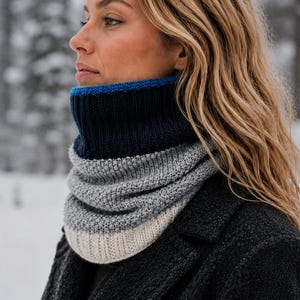 May include: A knit neck warmer featuring horizontal color-blocked stripes in navy blue, light gray, and cream. The top edge is finished with a thin bright blue band, while the navy and cream sections have a ribbed texture and the gray center is textured.