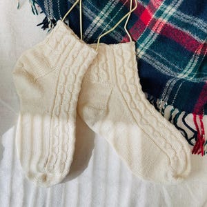 May include: A pair of cream-colored ankle socks featuring a vertical cable knit pattern along the sides and a ribbed texture on the cuffs. The socks are constructed with a soft knit fabric and include a gold-toned metal hanger for display.