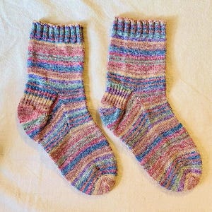 Op de afbeelding: Een paar handgebreide sokken met een meerkleurig garenpatroon met horizontale strepen in tinten roze, blauw, paars en geel. De sokken hebben een geribbelde boord aan de bovenkant, een gedefinieerd hielgedeelte en een afgeronde teen.