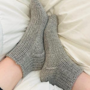May include: A pair of ankle-length socks knitted from light grey yarn. The socks feature a ribbed texture throughout the cuff and foot, with a distinct heel construction and a smooth, stockinette stitch pattern along the top of the foot area.