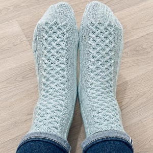 Puede incluir: Un par de calcetines de punto azul claro con un patrón de trenzas texturizado a lo largo del pie y la pierna, combinados con pantalones vaqueros azul oscuro. Los calcetines tienen un acabado de punto liso en la puntera y el talón.