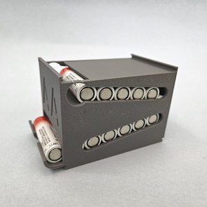 May include: A dark gray 3D-printed battery dispenser with the letters AA embossed on the side. The unit features two angled internal tracks designed to hold and dispense cylindrical AA batteries, with one battery resting in the front tray.