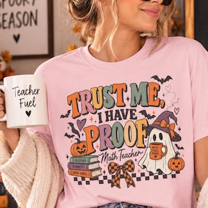 May include: A light pink t-shirt featuring a graphic with the text "TRUST ME, I HAVE PROOF" and "Math Teacher" alongside illustrations of a ghost wearing a witch hat, pumpkins, a stack of books, a checkered bow, and small bats.