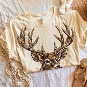 Puede incluir: Una camiseta de manga corta de color crema con un gran gráfico de una cabeza de ciervo con astas prominentes. Las astas y la cara del ciervo están rellenas con un patrón de camuflaje detallado de texturas forestales marrones, verdes y beige.