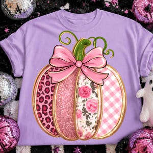 Könnte beinhalten: Ein hellviolettes T-Shirt mit einer großen Kürbisgrafik und einer rosa Schleife oben. Die Kürbissegmente zeigen verschiedene Muster, darunter rosa Leopardenmuster, rosa Glitzer, rosa Rosen auf weißem Grund und ein rosa Vichy-Karomuster.