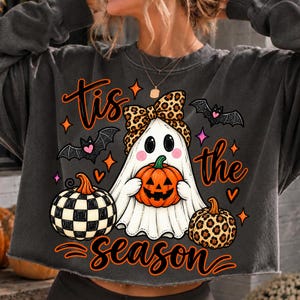 Può includere: Una felpa grigio antracite a maniche lunghe con la grafica di un fantasma bianco che indossa un fiocco leopardato e tiene in mano una zucca arancione. Il design include due pipistrelli, una zucca a scacchi e il testo 'tis the season'.