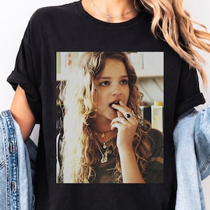May include: A black t-shirt featuring a rectangular graphic print of a person with long wavy blonde hair, wearing multiple necklaces and rings, with a finger near their mouth. The shirt has a crew neckline and short sleeves, shown in a black color.
