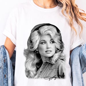 May include: A white t-shirt featuring a black and white graphic print of Dolly Parton with voluminous hair, wearing a denim shirt and hoop earrings. The print includes a small butterfly icon and the signature Dolly in cursive script at the bottom.