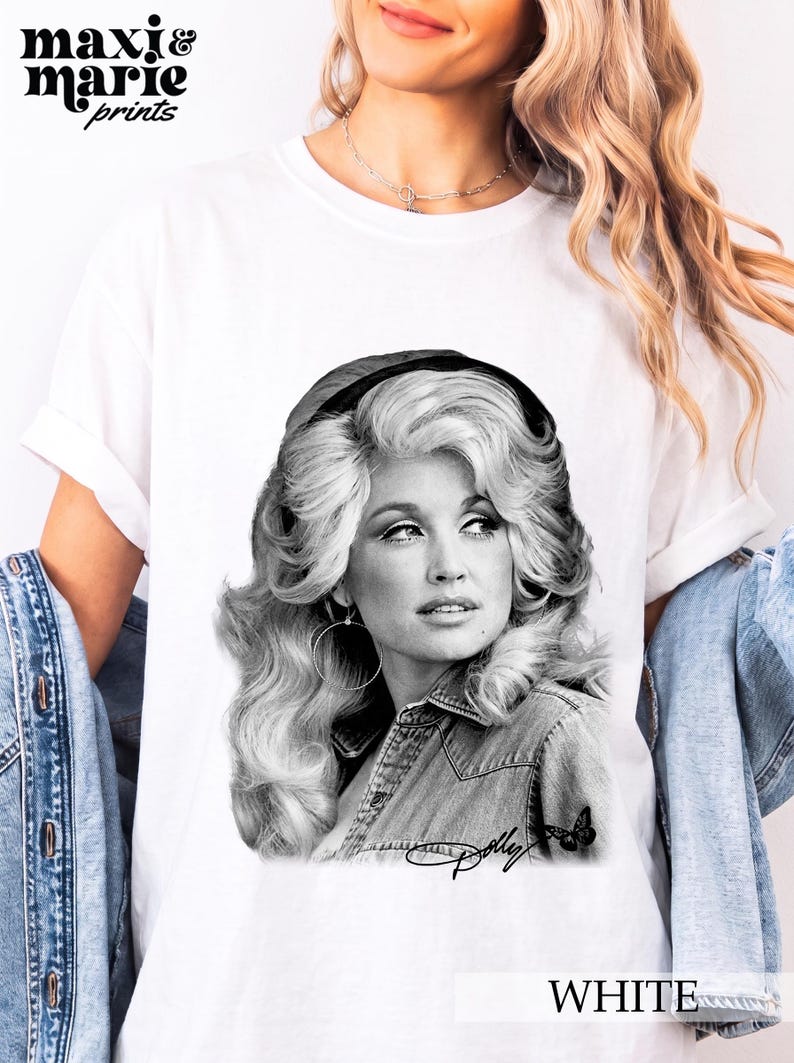 May include: A white t-shirt featuring a black and white graphic print of Dolly Parton with voluminous hair, wearing a denim shirt and hoop earrings. The print includes a small butterfly icon and the signature Dolly in cursive script at the bottom.