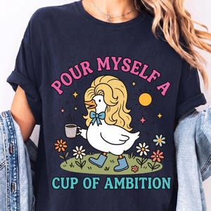 May include: A navy blue t-shirt featuring a graphic of a white duck with blonde hair, wearing blue boots and a blue bow, holding a coffee mug. The text reads "POUR MYSELF A CUP OF AMBITION" in pink and teal. The design includes flowers and a sun.