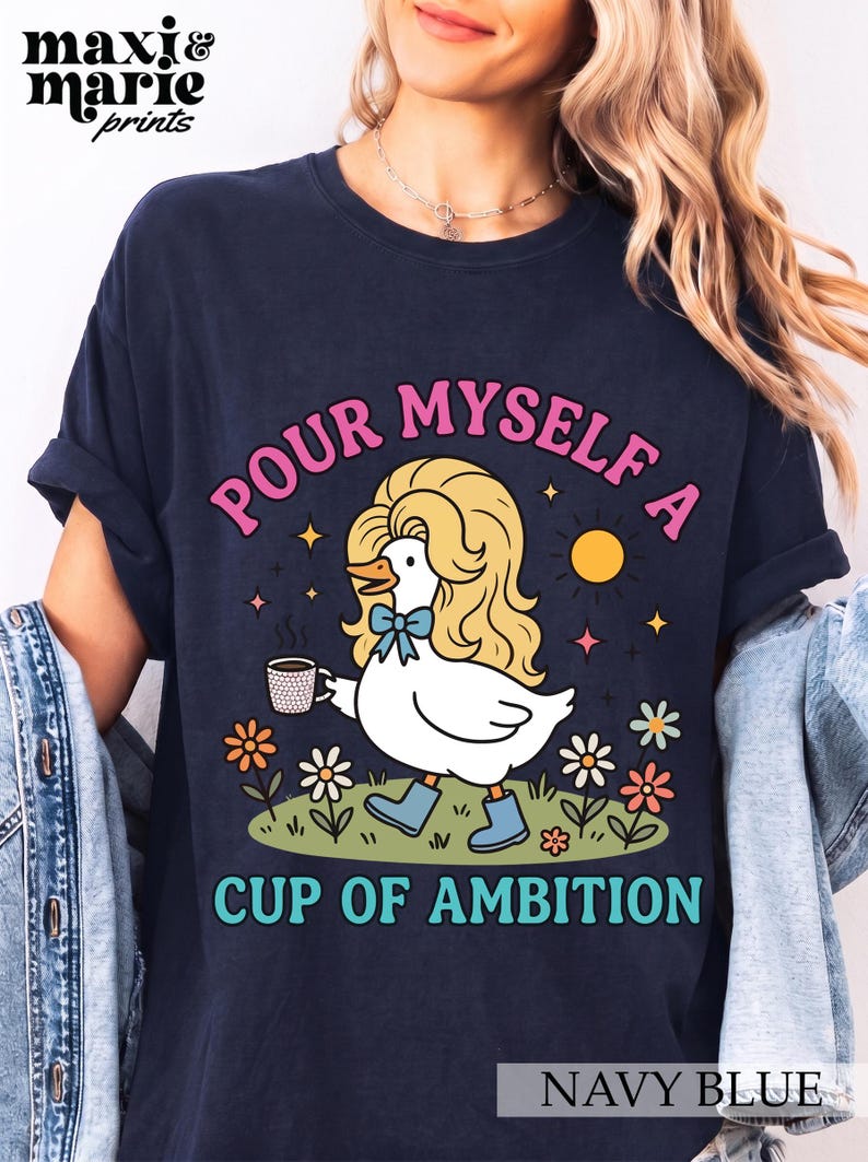 May include: A navy blue t-shirt featuring a graphic of a white duck with blonde hair, wearing blue boots and a blue bow, holding a coffee mug. The text reads "POUR MYSELF A CUP OF AMBITION" in pink and teal. The design includes flowers and a sun.