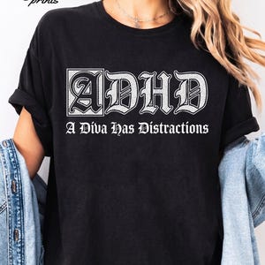 Könnte beinhalten: Ein schwarzes kurzärmeliges T-Shirt mit weißem Text, der ADHD und A Diva Has Distractions in einer gotischen Schriftart zeigt. Das T-Shirt hat einen Rundhalsausschnitt und eine lockere Passform. Der Markenname maxi & marie prints ist oben links sichtbar.