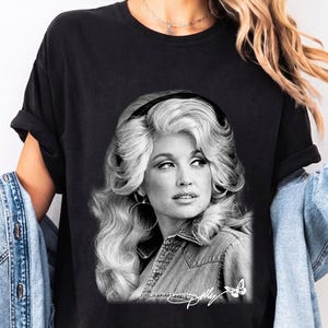 May include: A black t-shirt featuring a black and white photographic print of Dolly Parton with voluminous hair, wearing a denim shirt and hoop earrings. The graphic includes the signature Dolly and a small butterfly icon at the bottom right corner.