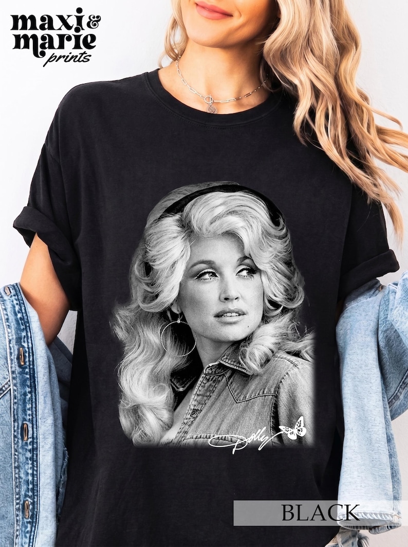 May include: A black t-shirt featuring a black and white photographic print of Dolly Parton with voluminous hair, wearing a denim shirt and hoop earrings. The graphic includes the signature Dolly and a small butterfly icon at the bottom right corner.