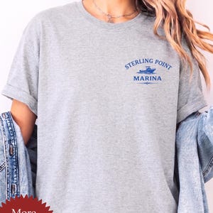 May include: A sports grey short-sleeve t-shirt featuring a blue graphic on the upper left chest. The graphic depicts a small boat above the text Sterling Point Marina. The shirt has a crew neckline and rolled sleeves, with a denim jacket draped over.
