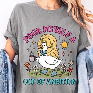 May include: A sports grey t-shirt featuring a graphic of a white duck with blonde hair, a blue bow, and blue boots holding a coffee cup. The text reads "POUR MYSELF A CUP OF AMBITION" in pink and blue letters, surrounded by flowers and a sun.
