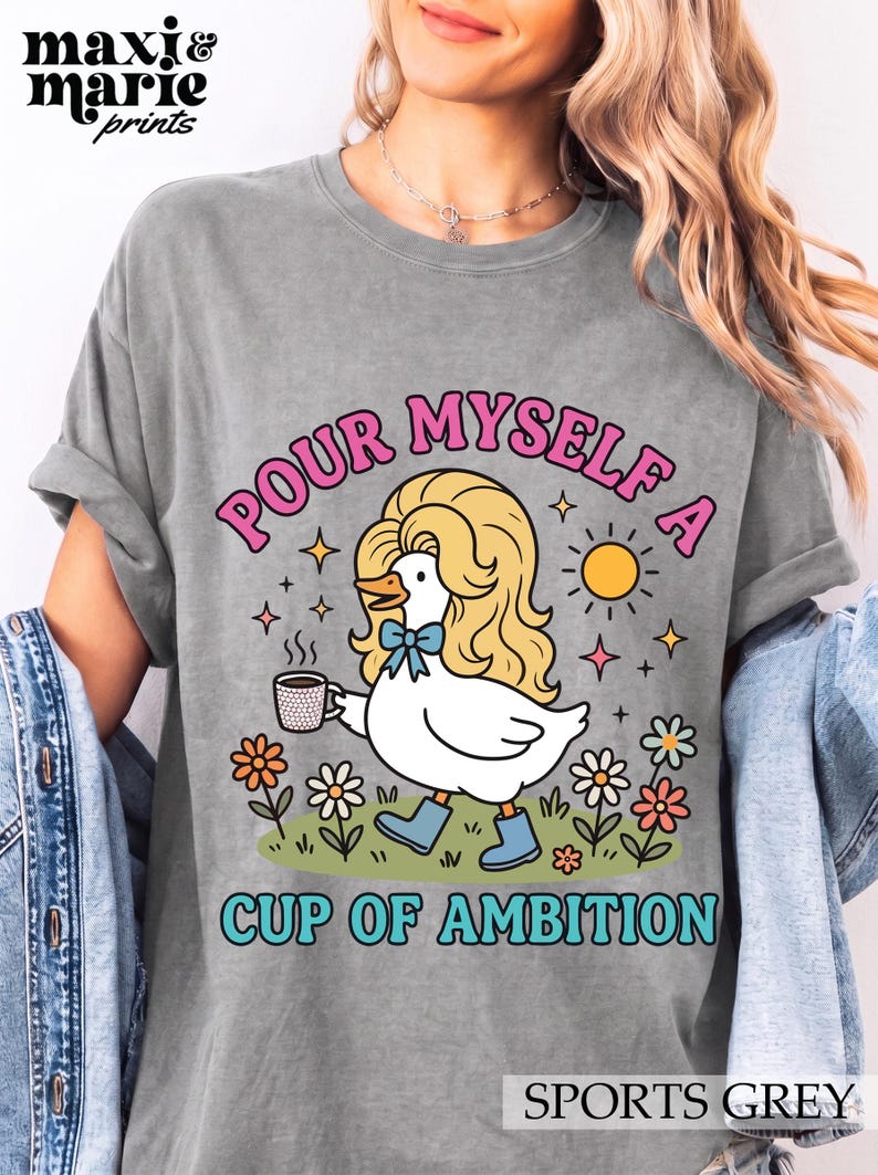 May include: A sports grey t-shirt featuring a graphic of a white duck with blonde hair, a blue bow, and blue boots holding a coffee cup. The text reads "POUR MYSELF A CUP OF AMBITION" in pink and blue letters, surrounded by flowers and a sun.