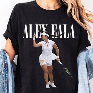 May include: A black short-sleeve t-shirt featuring the white text ALEX EALA above a graphic of a tennis player in a white outfit holding a racket. The top left corner displays the black text maxi & marie prints.