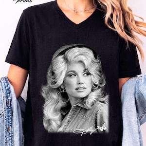 May include: A black V-neck t-shirt featuring a black and white photographic print of a woman with voluminous blonde hair wearing a denim shirt and hoop earrings. The graphic includes the signature Dolly and a small butterfly icon at the bottom.