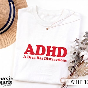 May include: A white t-shirt featuring the red text ADHD A Diva Has Distractions printed on the front. The shirt is displayed with a straw hat, a delicate necklace with a small star charm, and a bundle of dried eucalyptus and cotton stems.