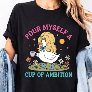 May include: A black t-shirt featuring a graphic of a white duck with blonde hair, a blue bow tie, and blue boots, holding a coffee mug. The text reads "POUR MYSELF A CUP OF AMBITION" in pink and teal, surrounded by colorful flowers and stars.
