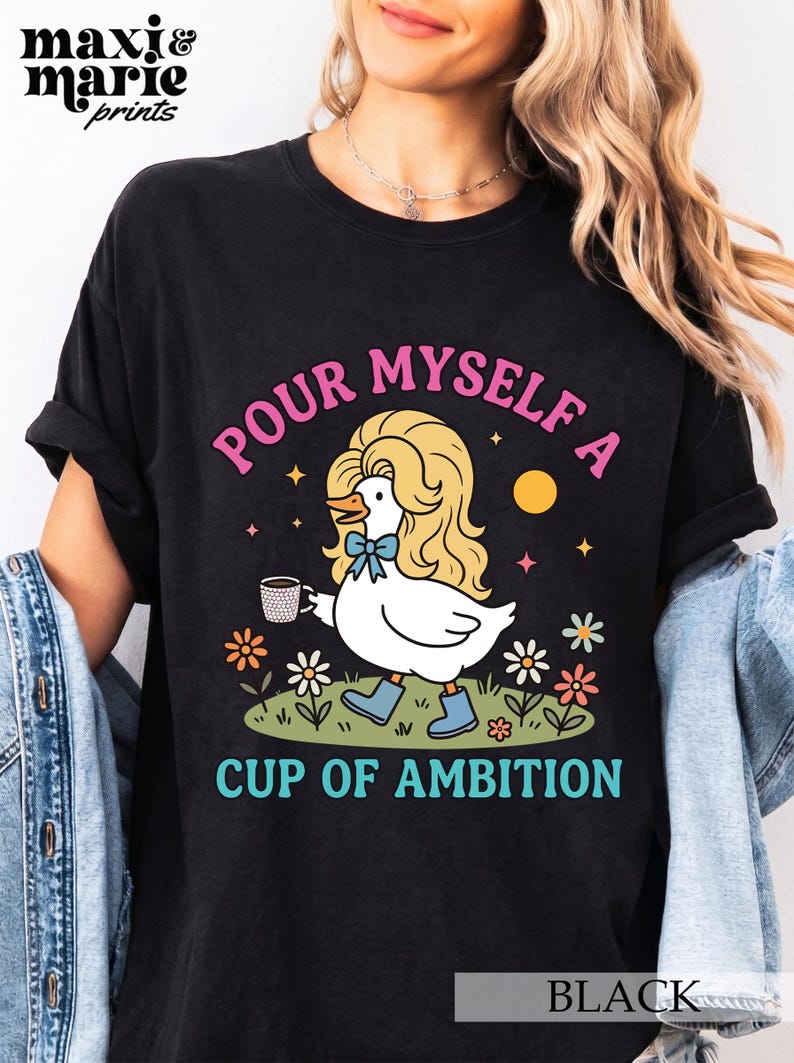 May include: A black t-shirt featuring a graphic of a white duck with blonde hair, a blue bow tie, and blue boots, holding a coffee mug. The text reads "POUR MYSELF A CUP OF AMBITION" in pink and teal, surrounded by colorful flowers and stars.