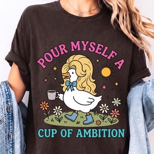 May include: A dark brown t-shirt featuring a graphic of a white duck with long blonde hair, wearing blue boots and a blue bow, holding a coffee mug. The text reads "POUR MYSELF A CUP OF AMBITION" in pink and teal, surrounded by flowers and a sun.