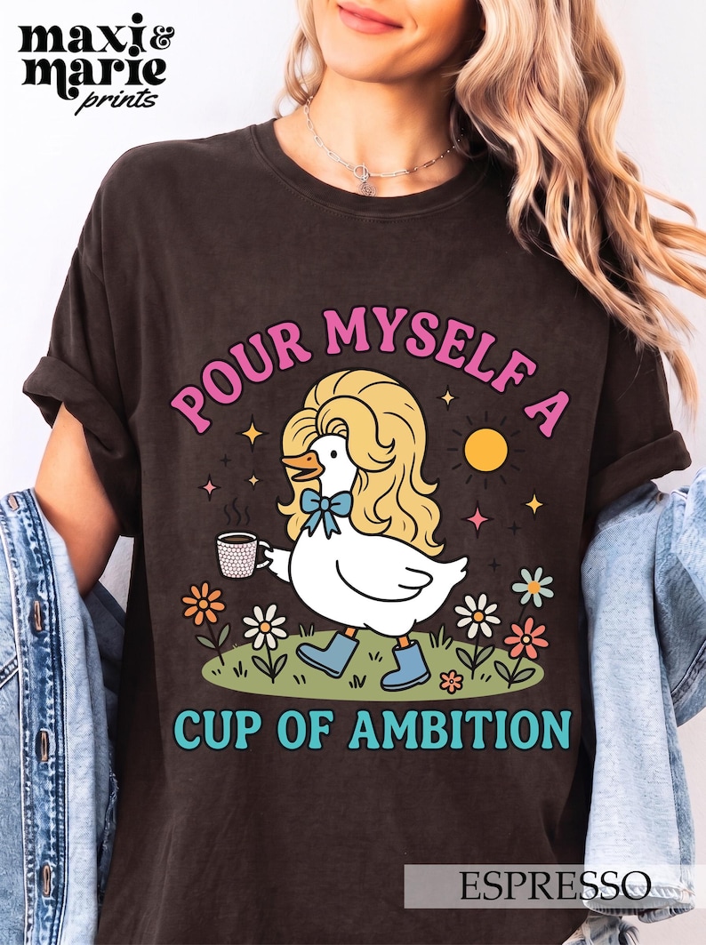 May include: A dark brown t-shirt featuring a graphic of a white duck with long blonde hair, wearing blue boots and a blue bow, holding a coffee mug. The text reads "POUR MYSELF A CUP OF AMBITION" in pink and teal, surrounded by flowers and a sun.