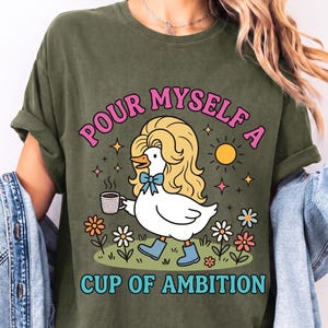 May include: A military green t-shirt featuring a graphic of a white duck with blonde hair, wearing blue boots and a blue bow, holding a coffee mug. The text reads "POUR MYSELF A CUP OF AMBITION" in pink and blue letters, surrounded by flowers and a sun.