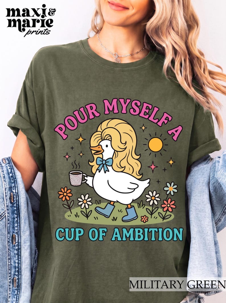 May include: A military green t-shirt featuring a graphic of a white duck with blonde hair, wearing blue boots and a blue bow, holding a coffee mug. The text reads "POUR MYSELF A CUP OF AMBITION" in pink and blue letters, surrounded by flowers and a sun.
