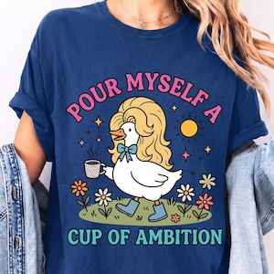 May include: A royal blue t-shirt featuring a graphic of a white duck with long blonde hair, wearing blue boots and a blue bow, holding a coffee mug. The text reads "POUR MYSELF A CUP OF AMBITION" in pink and teal letters, surrounded by flowers and a sun.