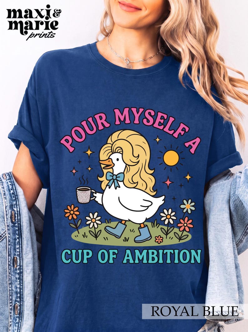 May include: A royal blue t-shirt featuring a graphic of a white duck with long blonde hair, wearing blue boots and a blue bow, holding a coffee mug. The text reads "POUR MYSELF A CUP OF AMBITION" in pink and teal letters, surrounded by flowers and a sun.