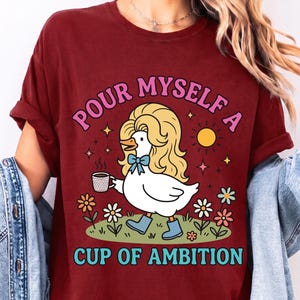 May include: A dark red t-shirt featuring a graphic of a white duck with blonde hair, wearing blue boots and a blue bow, holding a coffee mug. The text reads "POUR MYSELF A CUP OF AMBITION" in pink and blue letters, surrounded by flowers and a sun.