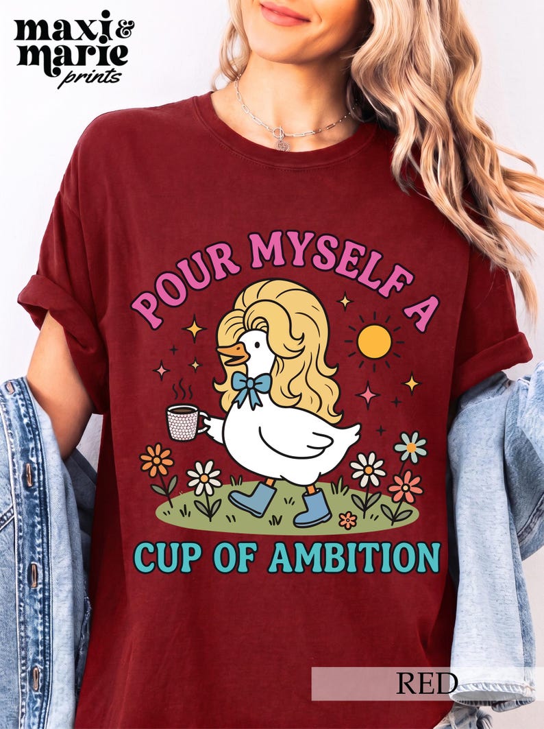 May include: A dark red t-shirt featuring a graphic of a white duck with blonde hair, wearing blue boots and a blue bow, holding a coffee mug. The text reads "POUR MYSELF A CUP OF AMBITION" in pink and blue letters, surrounded by flowers and a sun.