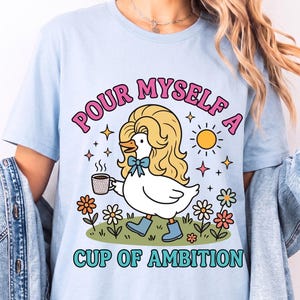 May include: A light blue t-shirt featuring a graphic of a white duck with blonde hair, wearing blue boots and a blue bow, holding a coffee mug. The text reads "POUR MYSELF A CUP OF AMBITION" in pink and teal letters, surrounded by flowers and a sun.