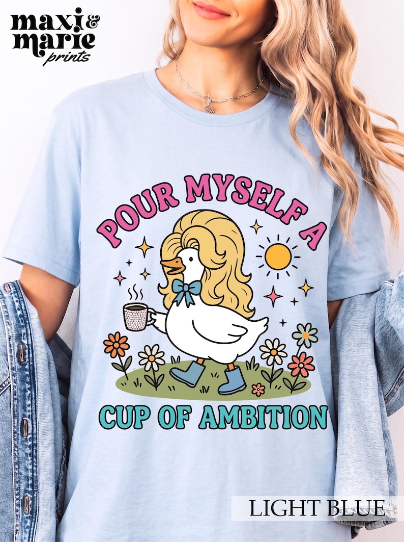 May include: A light blue t-shirt featuring a graphic of a white duck with blonde hair, wearing blue boots and a blue bow, holding a coffee mug. The text reads "POUR MYSELF A CUP OF AMBITION" in pink and teal letters, surrounded by flowers and a sun.