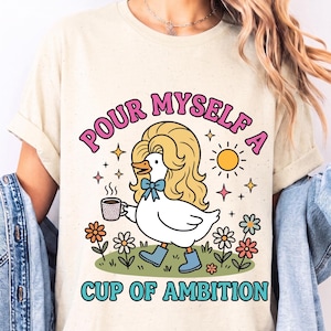May include: An ivory t-shirt featuring a graphic of a white duck with blonde hair, wearing blue boots and a blue bow, holding a steaming cup of coffee. The text reads "POUR MYSELF A CUP OF AMBITION" in pink and blue letters, surrounded by flowers and a sun.