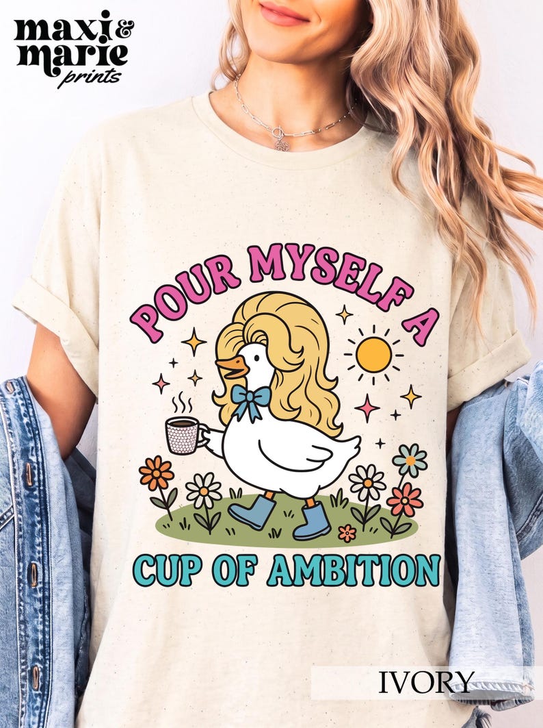 May include: An ivory t-shirt featuring a graphic of a white duck with blonde hair, wearing blue boots and a blue bow, holding a steaming cup of coffee. The text reads "POUR MYSELF A CUP OF AMBITION" in pink and blue letters, surrounded by flowers and a sun.
